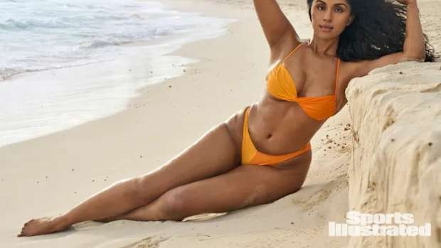 Manju lounges on the beach against a stone wall in an orange bikini,